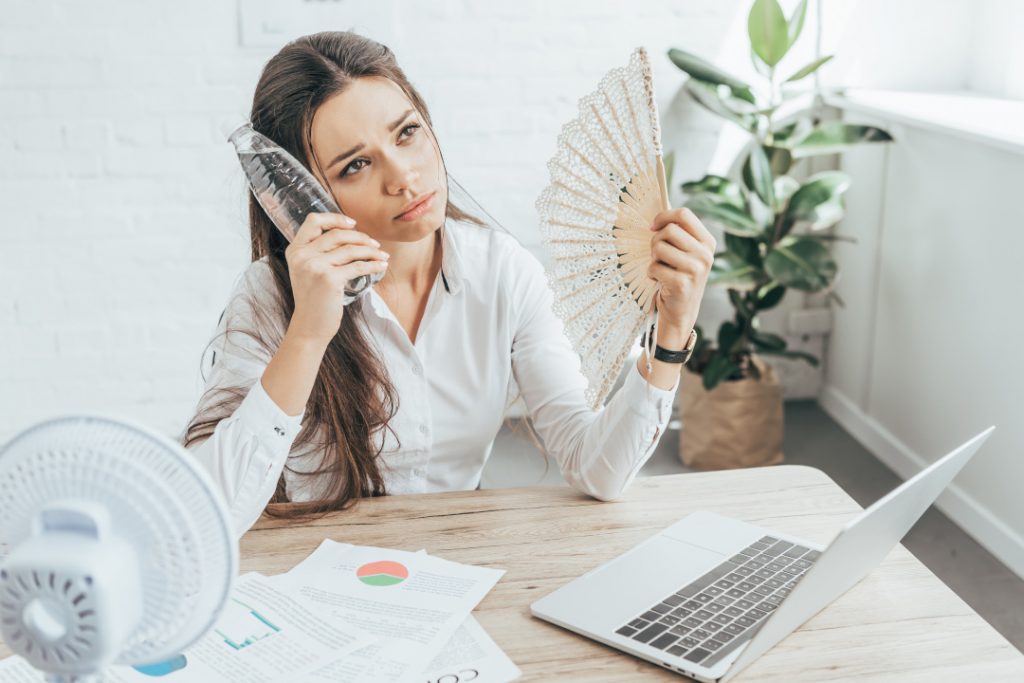 businesswoman cooling herself with electric fan, hand fan and bottle of water at workplace