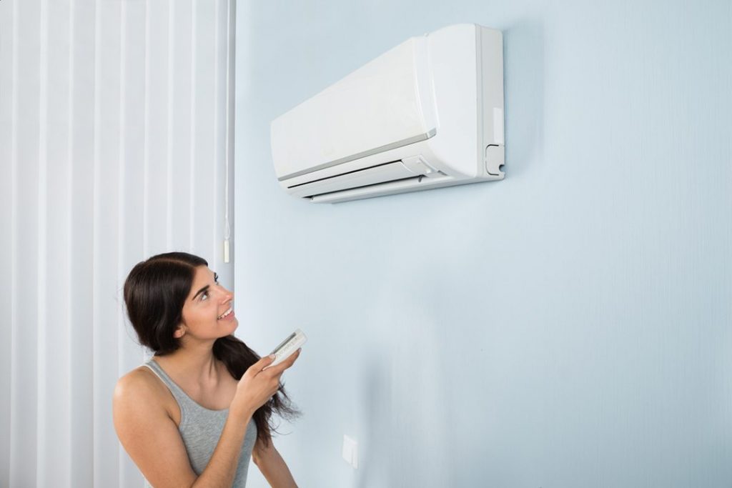 Young Woman Operating Air Conditioner With Remote Controller