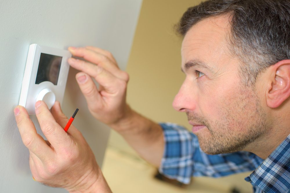 A man with a screwdriver inspects a wall-mounted thermostat