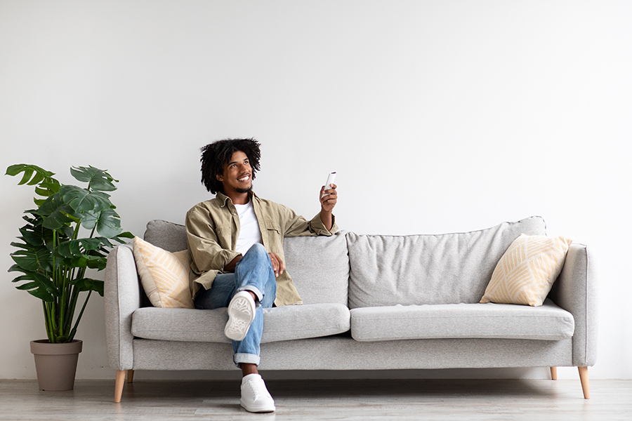 Happy Black Guy Sitting On Couch At Home, Operating Air Conditioner With Remote Controller, Smiling African American Man Adjusting Temperature Mode While Resting On Comfortable Sofa In Living Room